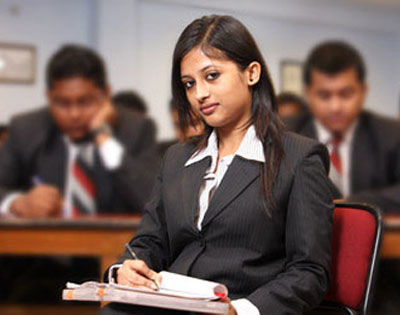 Female student taking notes during hotel management degree course class, surrounded by peers in formal uniforms.